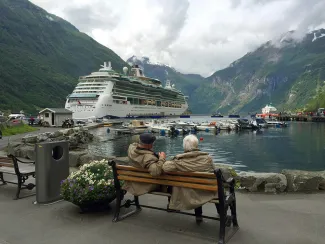 woman in brown coat sitting on brown wooden bench near white cruise ship during daytime by Julius Yls courtesy of Unsplash.